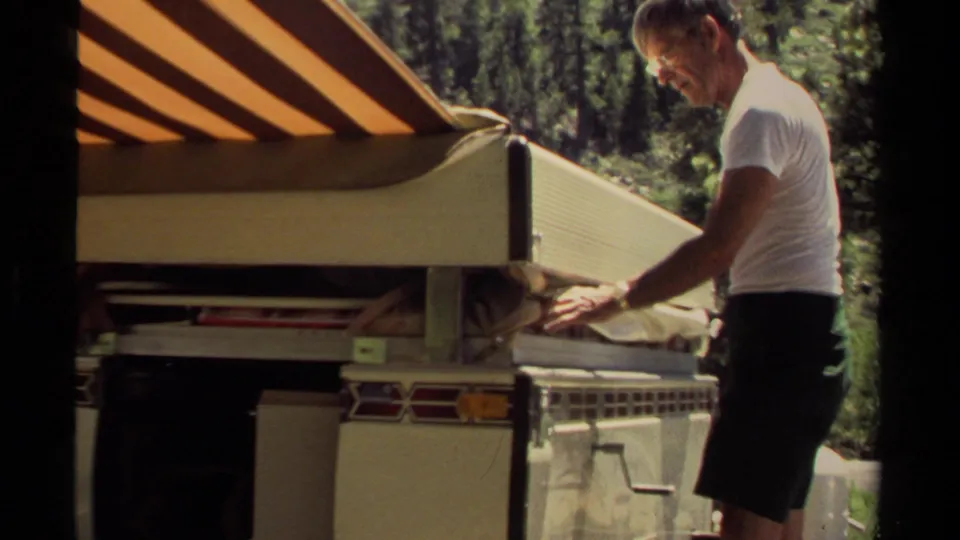 In Mendocino, California, 1980, a man in a white T-shirt works beside a tent trailer under trees while handling awning fabric.