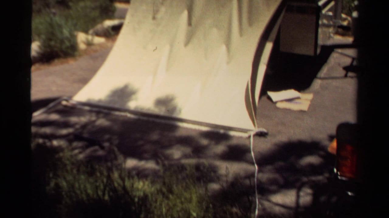 Partly folded tent trailer with pale canvas and campsite pad visible beside trees.