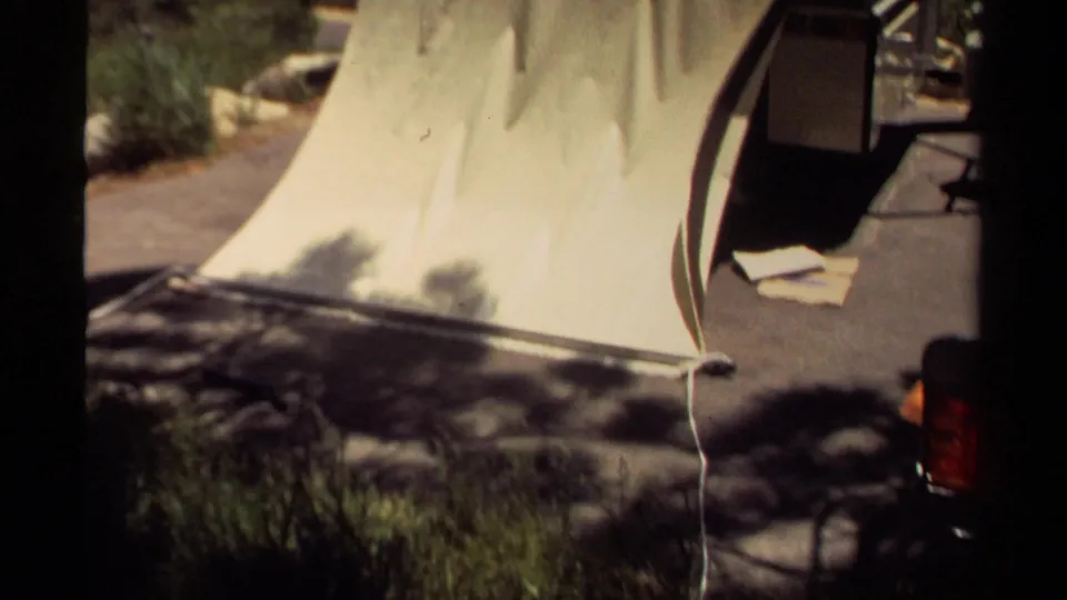 A pale canvas section of the 1980 Mendocino tent trailer lies partly folded on a paved campsite pad with trees and gear nearby.