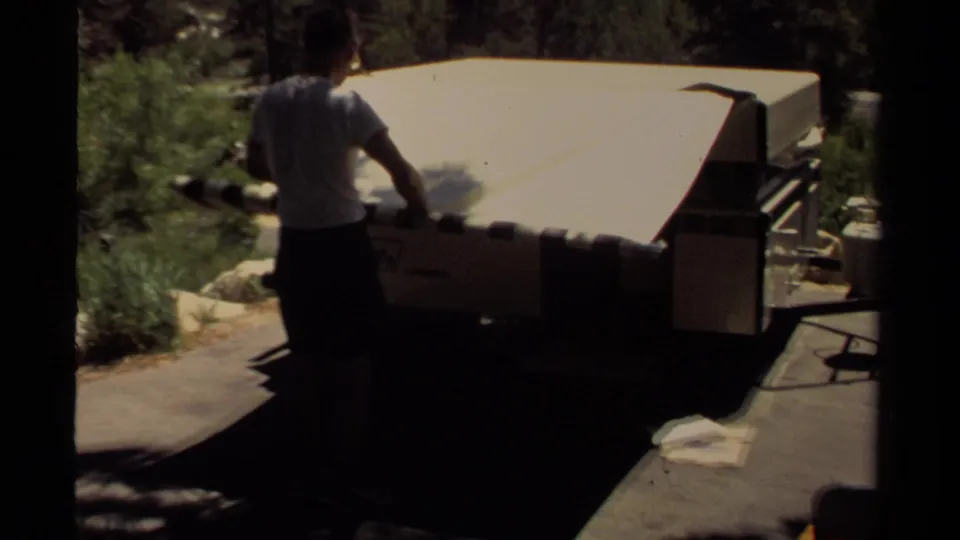 A man in shorts stands behind the tent trailer in Mendocino, California, as the pale canvas is folded down on the campsite pad.
