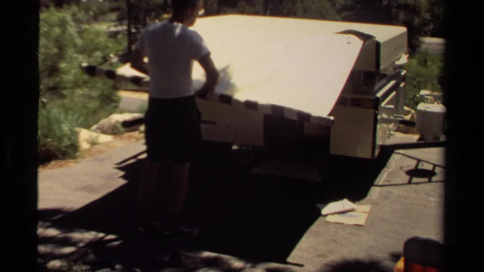 The man stands at the rear of the folded tent trailer in the 1980 Mendocino campsite scene, with a propane cylinder and trees nearby.