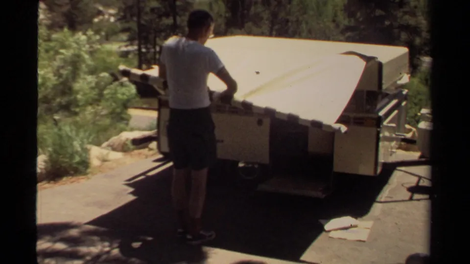 A man stands close to the rear of the tan camper, handling the folded top in a wooded Mendocino setting.