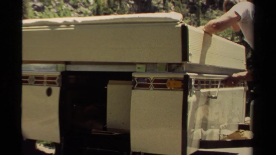 The tan pop-up camper fills the frame while the man continues securing the side canvas and panel in daylight.