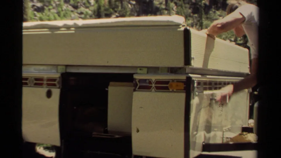 1980 Mendocino home-movie frame showing a white camper trailer side compartment being adjusted by a partially visible adult.