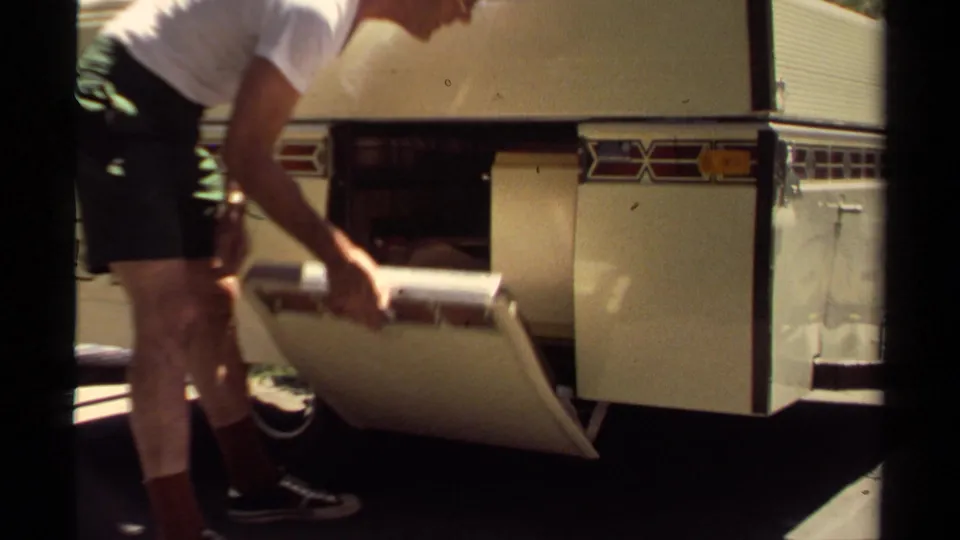 Adult man in casual clothing bends to close or guide a camper-trailer side compartment in 1980 Mendocino footage.