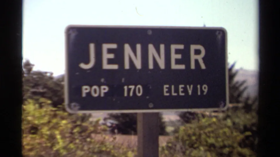 1980 8mm view of the Jenner, California town sign reading POP 170 ELEV 19, seen through a dark vertical window frame.