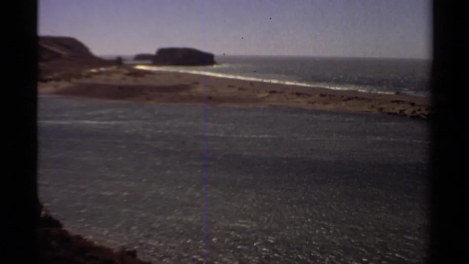 1980 8mm coastal view near the Jenner sequence, showing water, sandy shoreline, waves, and distant rocky outcrops through dark frame edges.