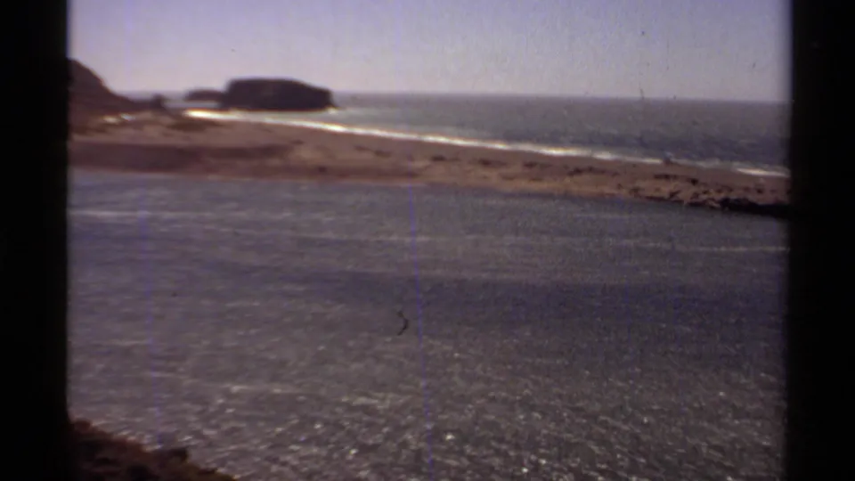 Soft 1980 8mm shoreline view from the Jenner-area clip, with ocean surface, pale surf, beach, distant rocks, and a dark window-frame edge.