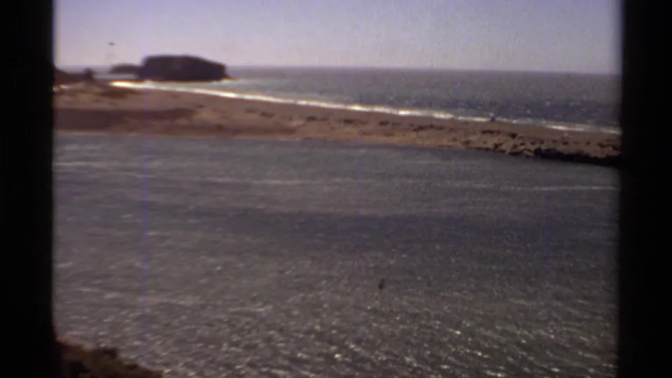 1980 8mm view of Mendocino coastal water, low surf, sandy shore, and a distant rocky headland.