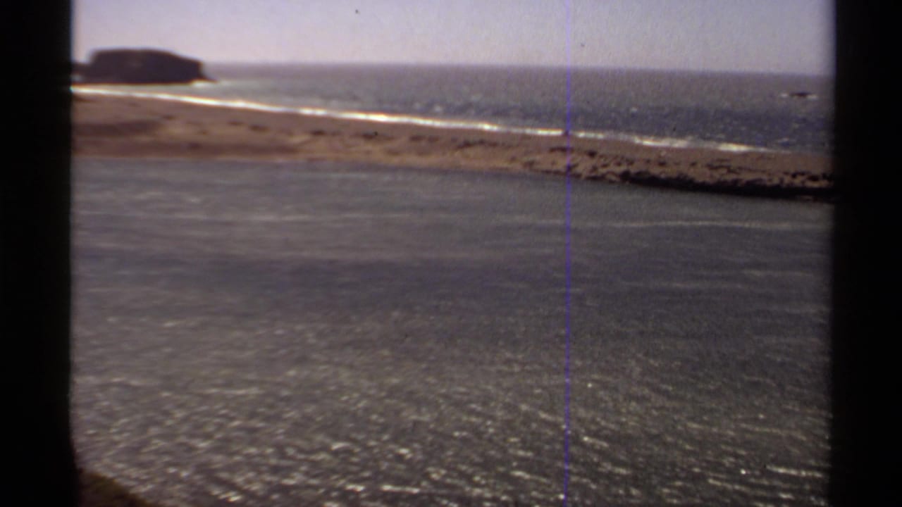 Ocean shoreline with breaking waves, distant rocks, and dark film borders.