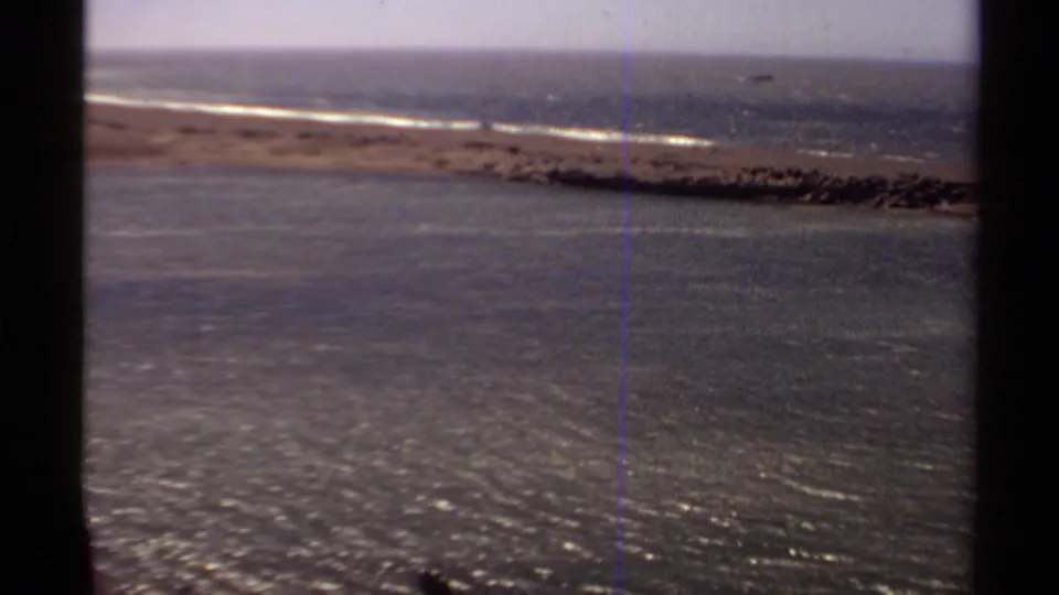 1980 8mm coastal view from Mendocino, showing rippled water in front of sand, surf, and a low rocky shoreline.