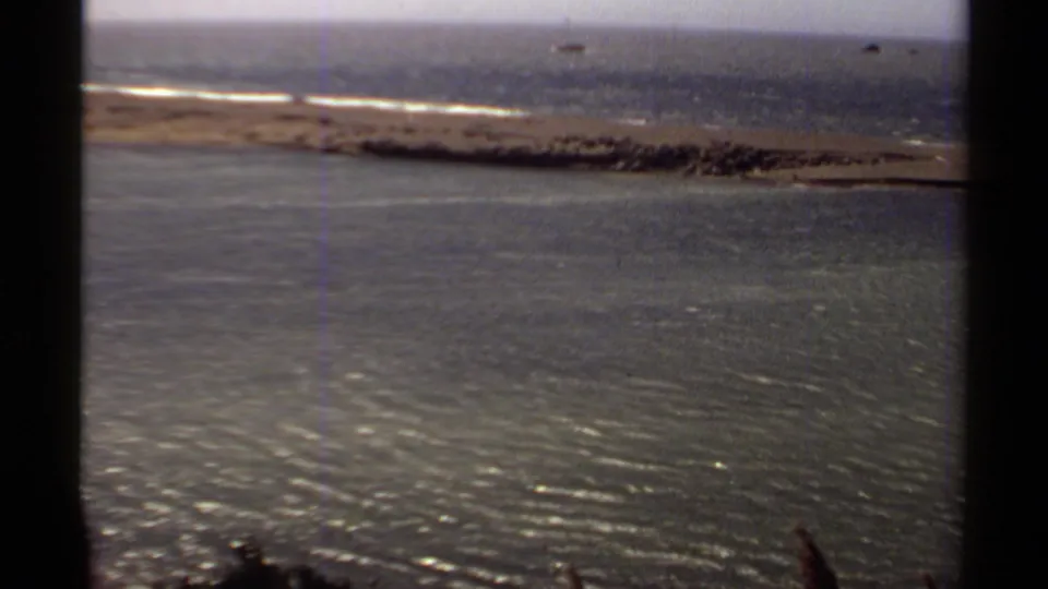 Mendocino coastal home-movie frame with shimmering water, a sandy shoreline, low surf, and small distant rocks offshore.