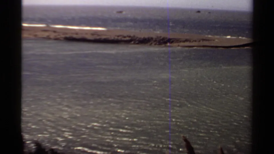 1980 Mendocino coastal home-movie frame showing open water, a narrow sandbar, small surf lines, and dark foreground framing.