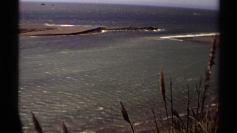 1980 Mendocino coastal frame with wind-rippled water, a sandbar, gentle wave breaks, and silhouetted shoreline grasses.