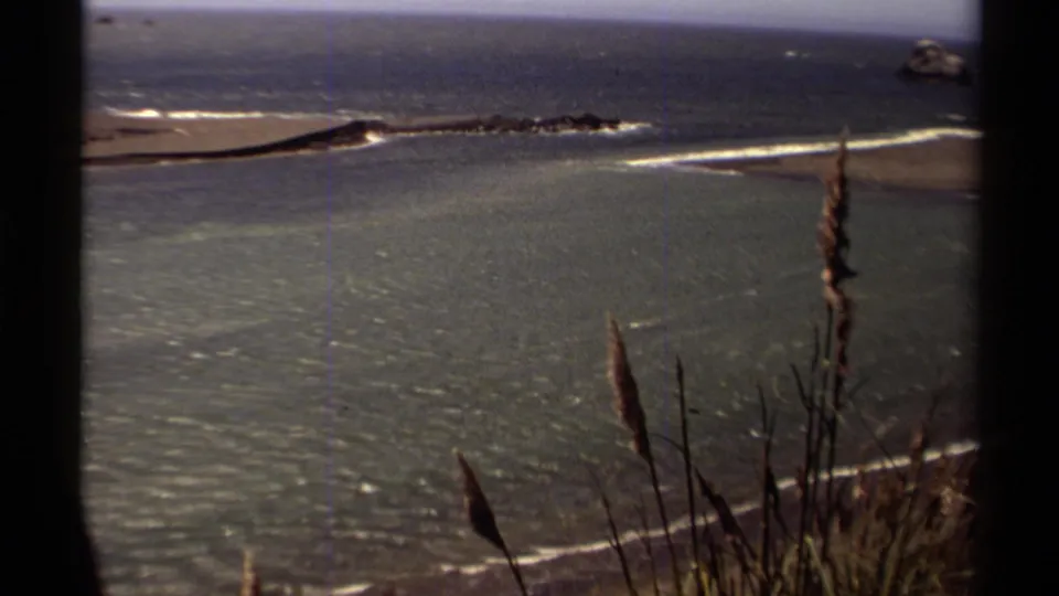Mendocino coastal view from 1980 8mm footage, with a sandy beach, surf lines, offshore rocks, and tall grasses in the foreground.
