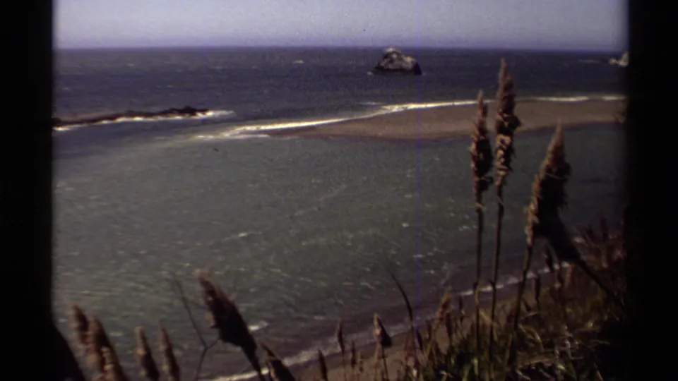 1980 8mm view of the Mendocino coast, showing surf across a sandbar, offshore sea stacks, and tall grasses near the camera.