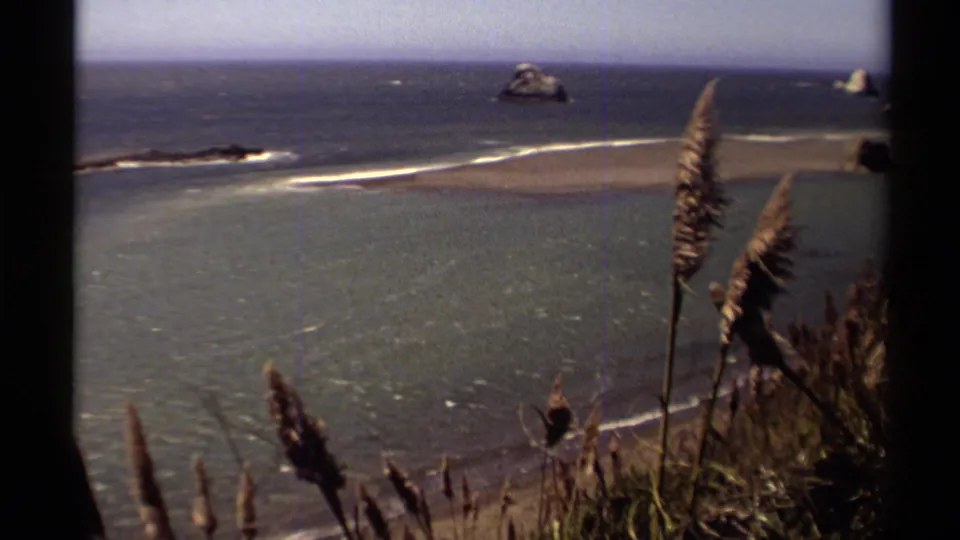 Vintage 8mm Mendocino shoreline frame with a sandy bar, offshore rocks, rippled water, and windblown grasses in front.