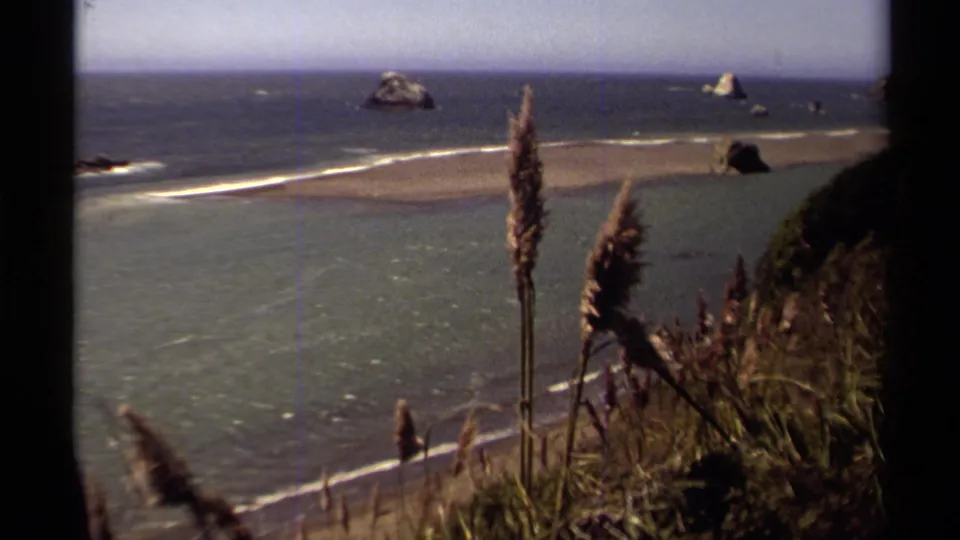 Mendocino, California coastal scene from 1980 8mm footage, with surf, sand, offshore sea stacks, and grasses along the cliff edge.