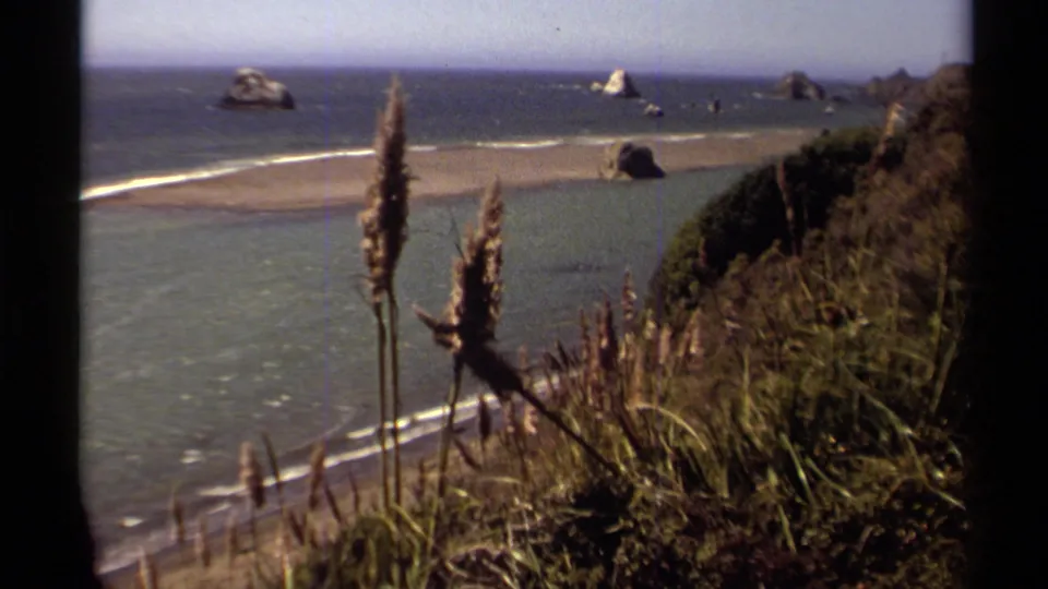 A vintage 8mm view of the Mendocino shoreline, with tall reeds and cliff vegetation above beach, waves and offshore sea stacks.