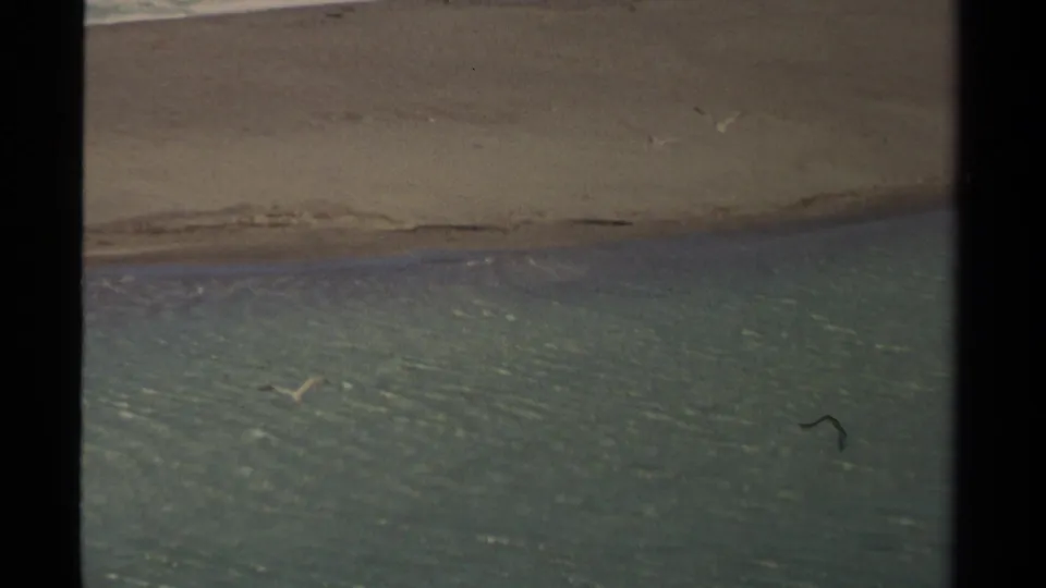 A sandy coastal spit crosses the frame with teal water on both sides, filmed in soft 1980 8mm Mendocino footage.