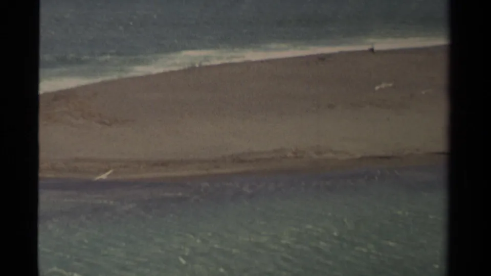 The Mendocino coastal scene shows a sandy shoreline between bands of water, with tiny distant marks near the surf and visible 8mm grain.