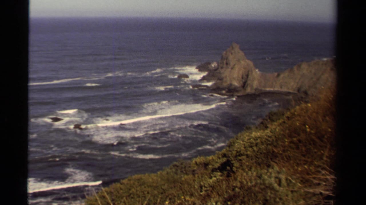 1980 Mendocino coastal view with Pacific waves, a rocky sea stack, cliff vegetation, and visible 8mm frame borders.