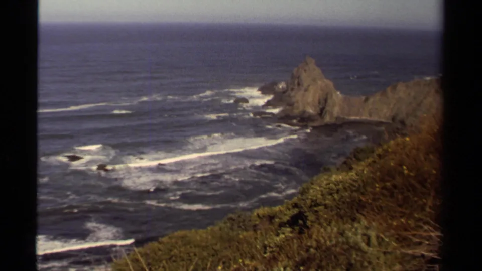 1980 Mendocino coastal view with Pacific waves, a rocky sea stack, cliff vegetation, and visible 8mm frame borders.