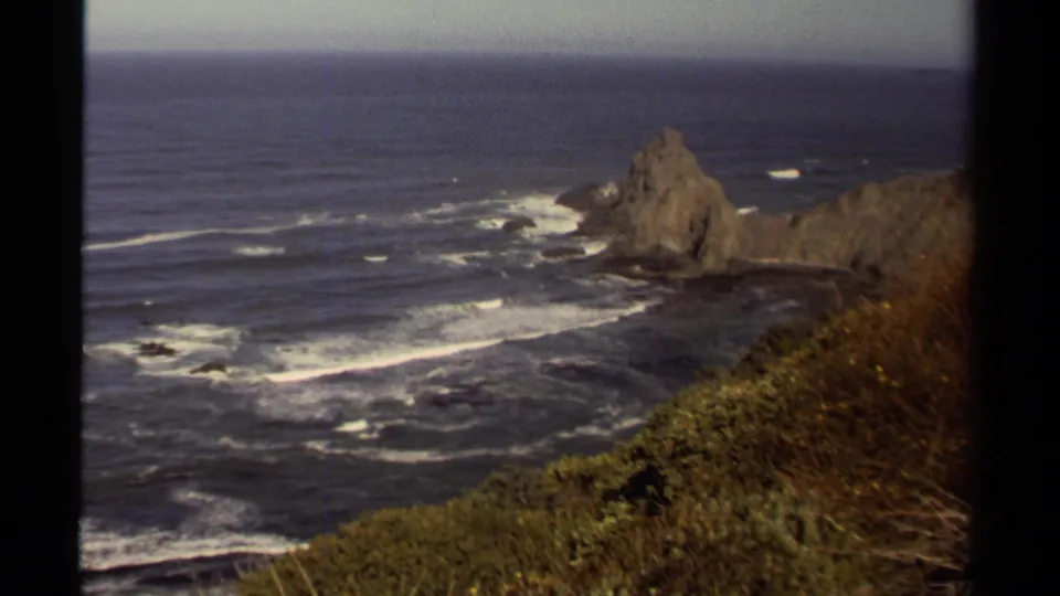 1980 Mendocino home-movie frame looking over cliffside vegetation toward surf and a rocky sea stack in the Pacific.