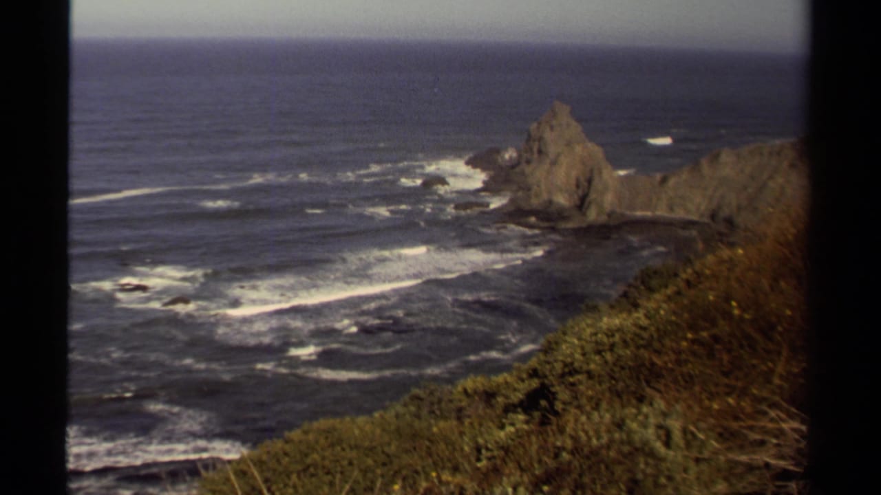 1980 8mm view of the Mendocino coast, showing a sea stack, dark ocean water, surf lines, and vegetation along a cliff edge.