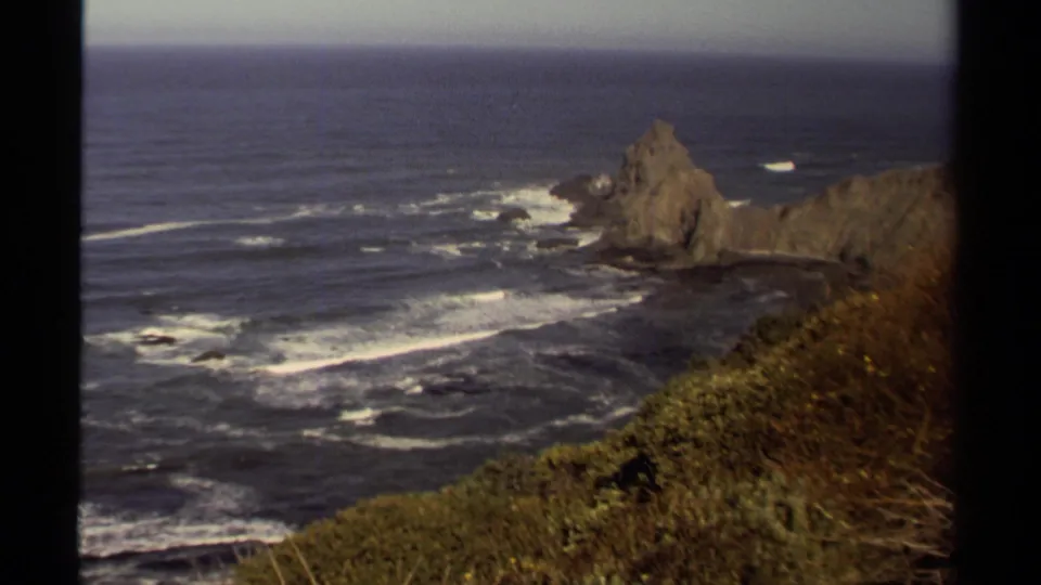 1980 8mm view of the Mendocino coast, showing a sea stack, dark ocean water, surf lines, and vegetation along a cliff edge.