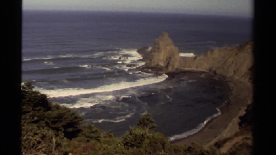 1980 8mm coastal view in Mendocino, showing a secluded dark sand cove, white wave lines, rocky headland, and foreground coastal plants.