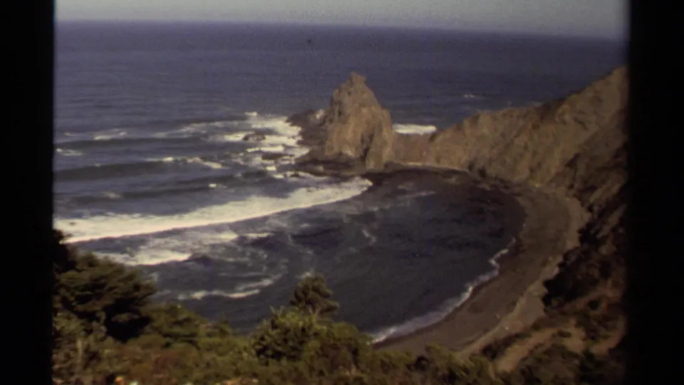 Vintage Mendocino 8mm frame showing a broad view of the cove, foamy surf, rugged cliff walls, and the sea stack beyond the shoreline.