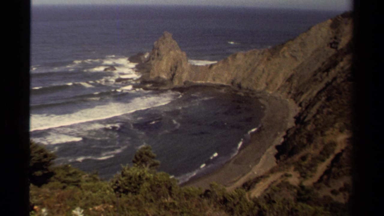 Coastal cove seen from above with cliffs, sea stack, surf, and beach.