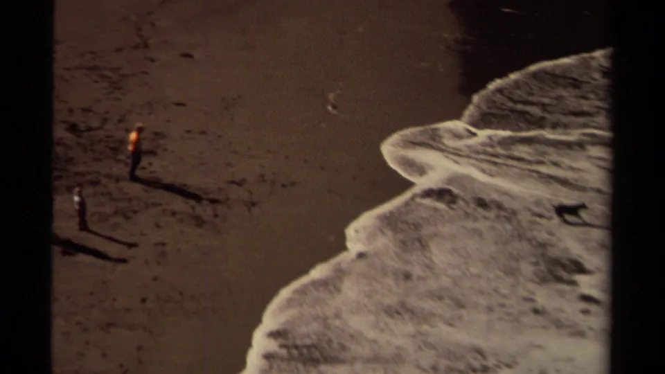 Mendocino coastal home-movie frame from 1980 showing two distant beachgoers near white surf on dark sand.