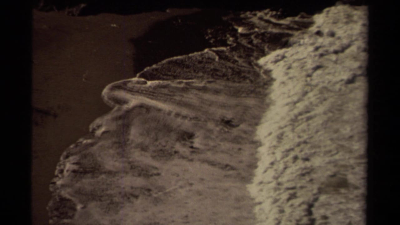 Foamy wave line curves across dark sand with a rocky coastal edge nearby.