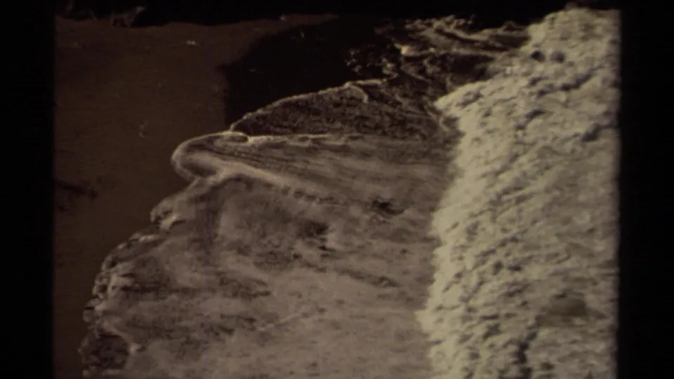 1980 Mendocino coast frame showing white ocean foam spreading over dark beach sand near a rocky cove edge.