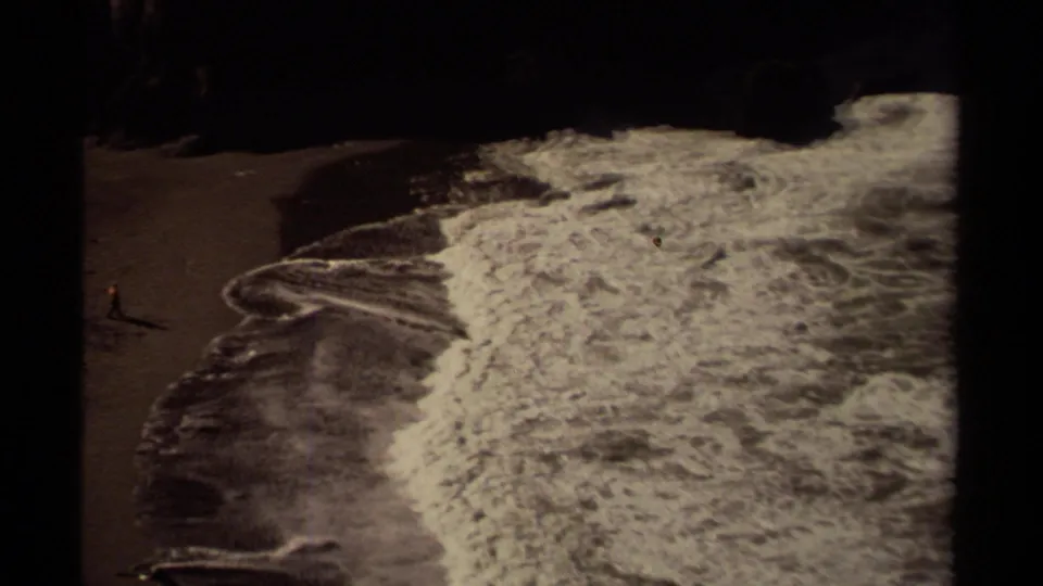 Mendocino 8mm frame from 1980 with a small beachgoer near the shoreline and white surf under a dark cliff edge.