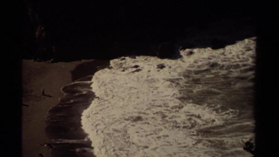 1980 Mendocino shoreline frame showing foamy surf sweeping across dark sand near a shadowed rocky cove.