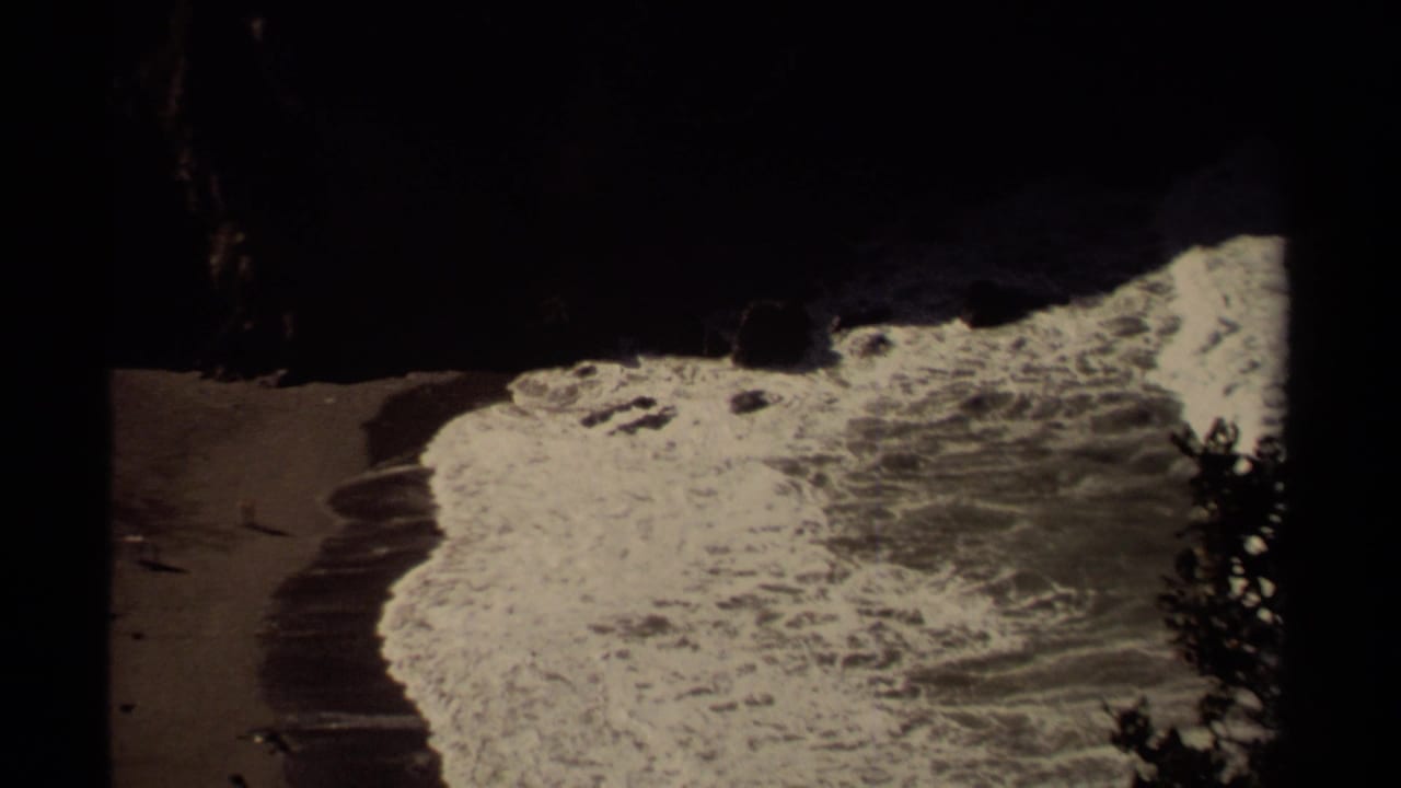 1980 8mm Mendocino coast frame showing surf washing onto a sandy beach beneath dark rock formations.