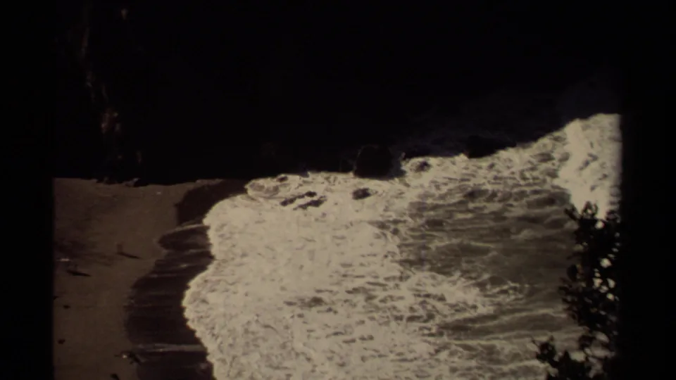 1980 8mm Mendocino coast frame showing surf washing onto a sandy beach beneath dark rock formations.