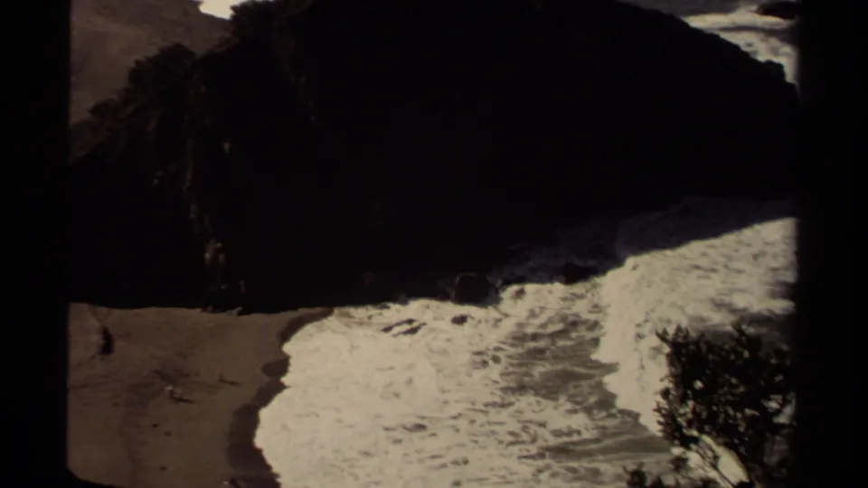 Vintage 8mm view of Mendocino, California surf wrapping around a dark coastal rock and sandy shoreline.