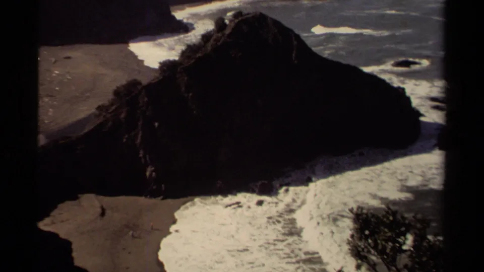 1980 Mendocino beach footage frame with a shadowed bluff, sandy cove, breaking waves, and tiny beachgoers below.
