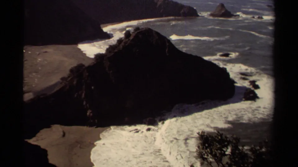 Mendocino 1980 8mm frame looking over a dark coastal rock toward foamy surf, beach sand, and offshore rocks.