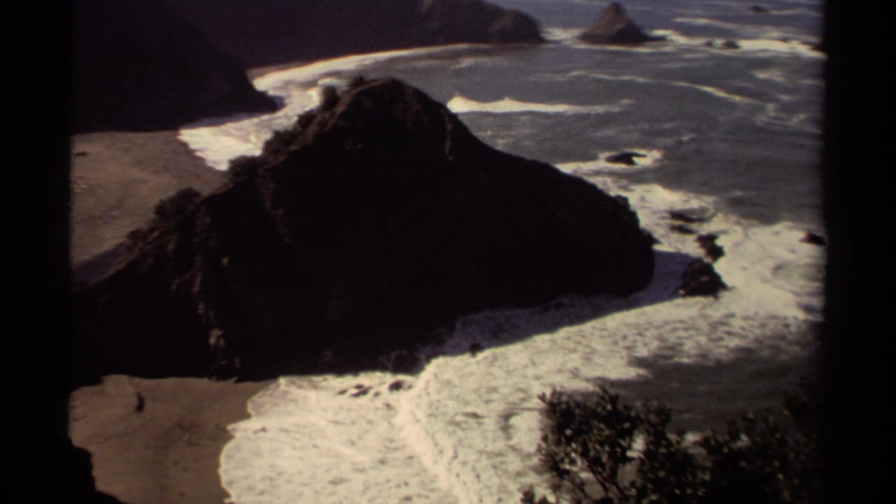 Rocky shoreline seen from above with waves breaking around a dark headland and sandy beach.