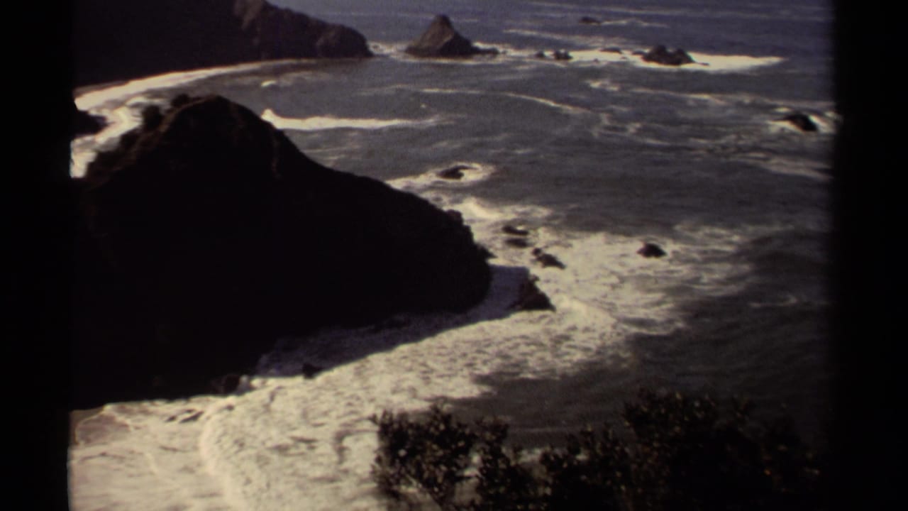 1980 Mendocino shoreline view with a dark coastal rock mass, foamy surf, blue ocean, and scattered sea stacks offshore.