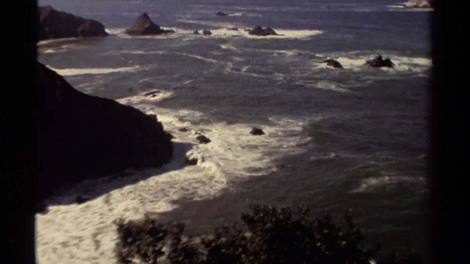 1980 Mendocino coast frame showing surf patterns below an elevated viewpoint, with a cliff edge, offshore rocks, and blue water.