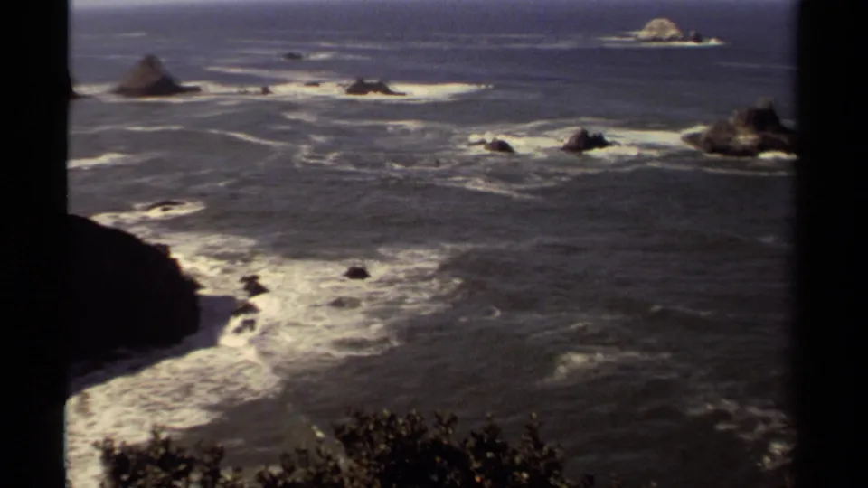 1980 Mendocino home-movie frame with a wider Pacific view, white surf, dark rock outcrops, sea stacks, and brush at the bottom edge.