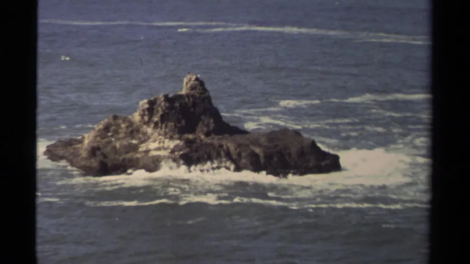 Closer 8mm frame of a large offshore rock near Mendocino, with waves breaking around its base.
