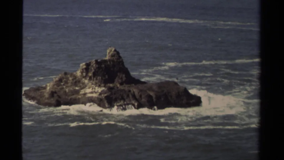 1980 Mendocino coastal home-movie frame showing a large sea rock, surrounding surf, and muted blue-gray ocean water.