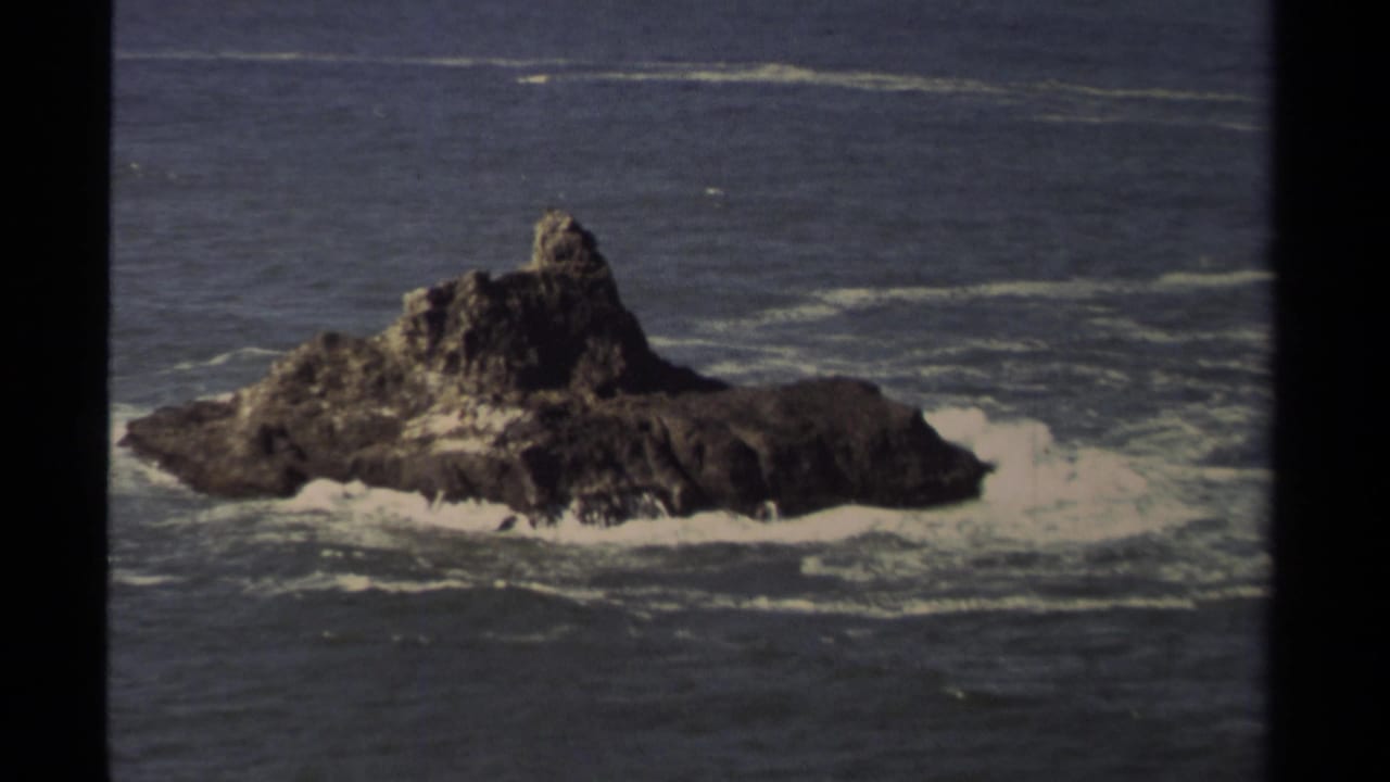 1980 8mm Mendocino coastal view of a rugged offshore rock with white surf breaking around its base.